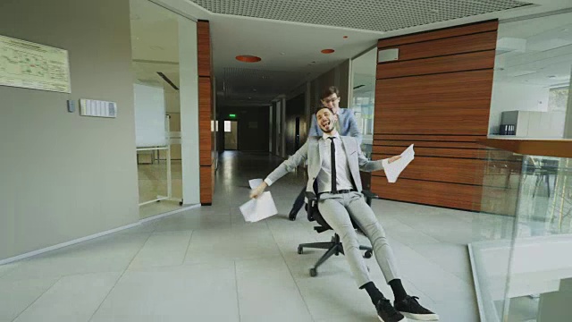 杜利拍摄了两个疯狂的商人在现代商业中心的大厅里，坐在办公椅上，把文件扔在地上玩耍视频素材