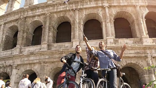 三个快乐的年轻朋友游客与自行车和背包在罗马斗牛场自拍与智能手机和木棍有乐趣在阳光明媚的日子慢动作稳定相机视频素材