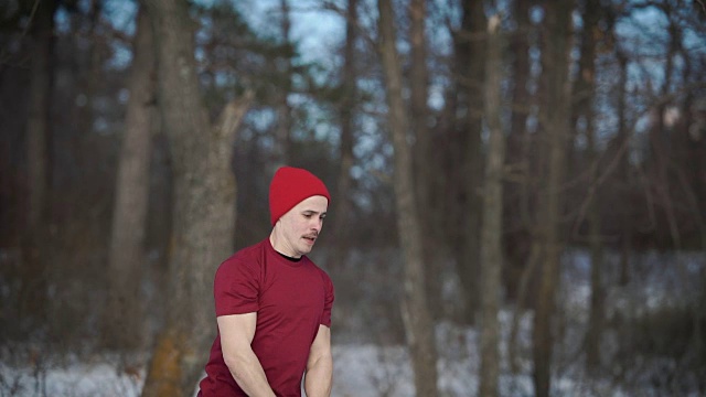 一个戴着红帽子的举重运动员在冬天的森林里举起和放下一个沉重的壶铃视频素材