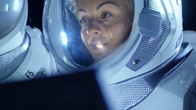 穿着太空服的女宇航员在笔记本电脑上工作，探索新发现的行星，与地球交流。背景中是她的船员和居住地。外星殖民的概念。视频素材