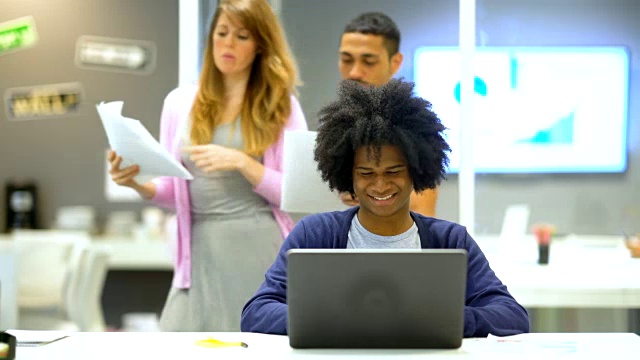 一位非裔美国设计师在他的笔记本电脑上工作得很开心，办公室里的人从他身边走过视频素材