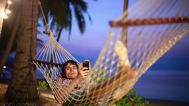 年轻英俊的男人在海滩的吊床上玩智能手机。异国情调的岛屿在暮色中与美丽的颜色的天空。1920 x1080视频素材