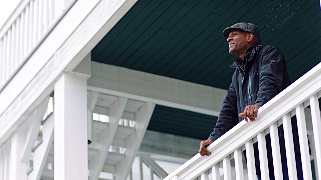 一名身穿夹克的非裔美国男子斜倚在户外阳台的栏杆上，微笑着互相搓着手欣赏风景。视频素材