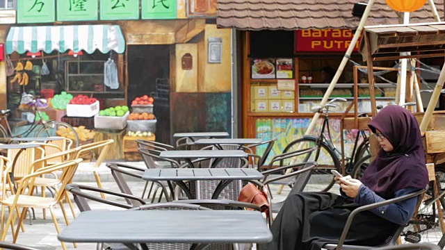 美丽的东南亚穆斯林妇女戴着头巾在户外咖啡馆使用智能手机的肖像，有三种色调(蓝-橙，对比和原始)视频下载