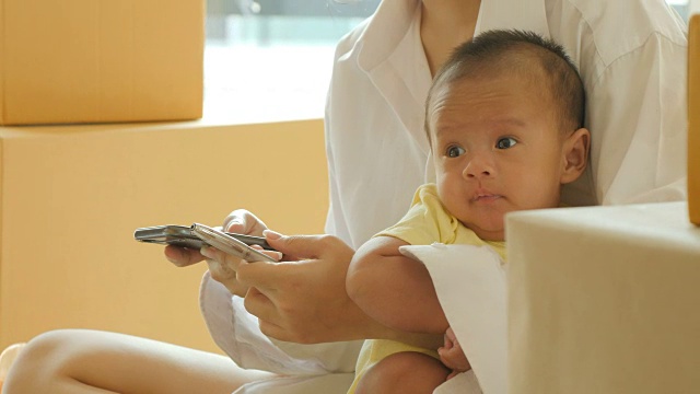 年轻的亚洲母亲抱着婴儿，检查智能手机上的客户订单和在线送货，准备在卧室打包。视频下载