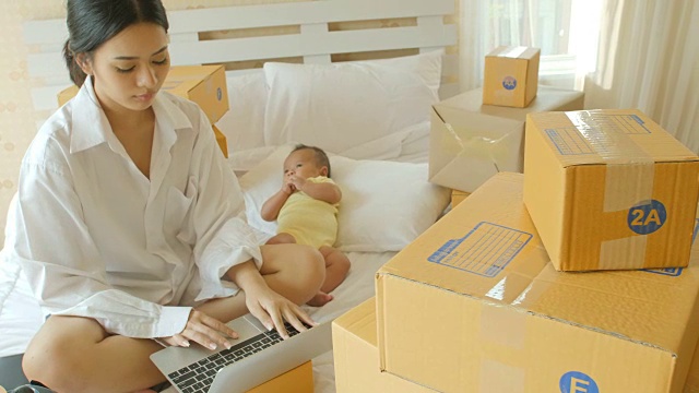 年轻的亚洲母亲和她的新生儿检查订单从笔记本电脑客户和在线交付准备装箱在卧室。视频下载