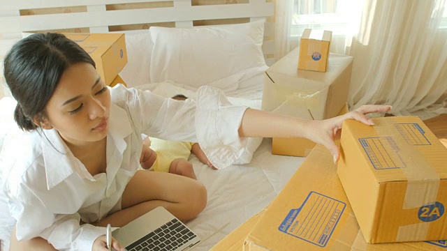 年轻的亚洲母亲和她的新生儿检查订单从笔记本电脑客户和在线交付准备装箱在卧室。视频购买