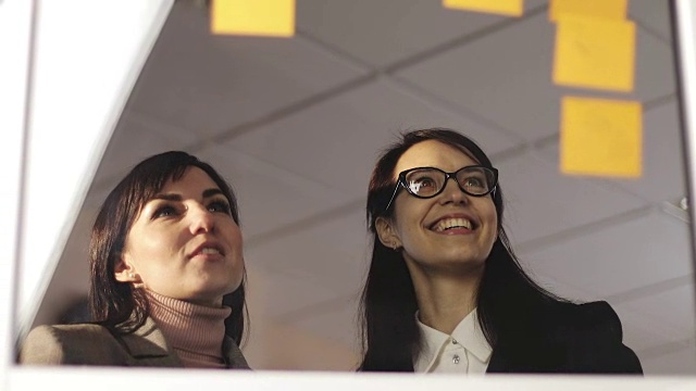 中年商业妇女和年轻的女孩看玻璃上的贴纸在商务会议和工作讨论在办公室空间。视频素材