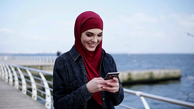 戴着头巾的迷人年轻女孩在微笑着给某人发短信，在智能手机上滚动着什么东西。户外慢镜头视频下载