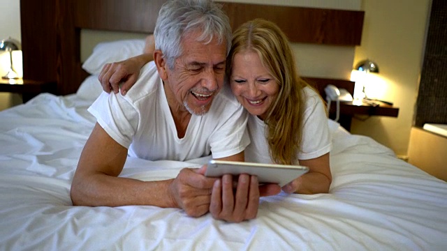 一对快乐的老年夫妇躺在床上，在平板电脑上看照片，开心地笑着视频素材