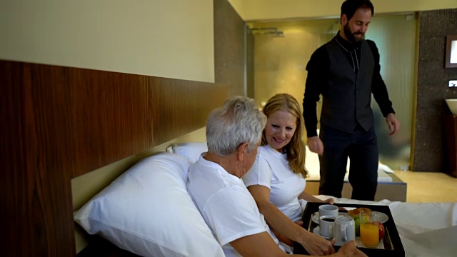 一对老年夫妇在一个浪漫的度假胜地躺下，一个男人在他们的酒店房间的床上给他们端来早餐视频素材