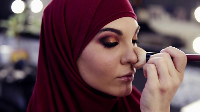 这个美丽的女孩用传统的阿拉伯紫色雪纺头巾遮住了她的头部，在她的脸颊上涂上了难以辨认的化妆师的手。视频下载