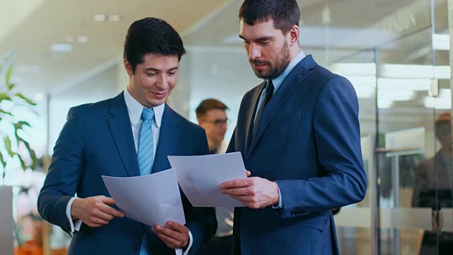 两个商人在公司大楼走廊握手，他们比较文件。忙碌的办公室生活和许多忙碌的商务人员。视频素材