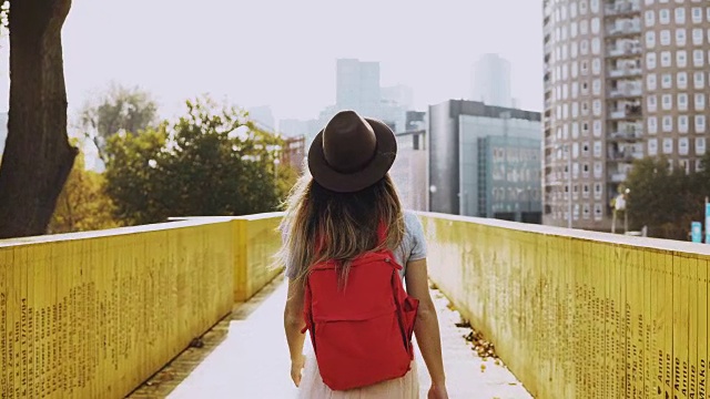 戴着帽子长发的女孩走在城市的桥上。一个背着红色背包站在黄色墙壁中间的白人女人。后视图4 k视频素材