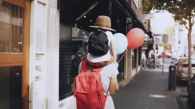 母亲背着儿子沿街走。女孩带着一个戴着帽子和两个气球的快乐小孩走着。4 k视频素材