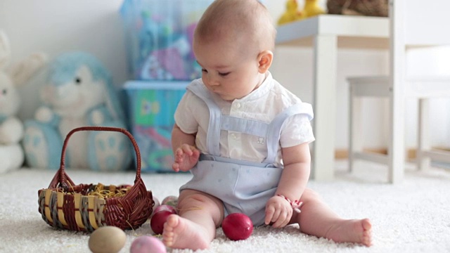 可爱的蹒跚学步的小男孩，在阳光明媚的客厅里玩着复活节巧克力兔子和五颜六色的复活节彩蛋视频下载