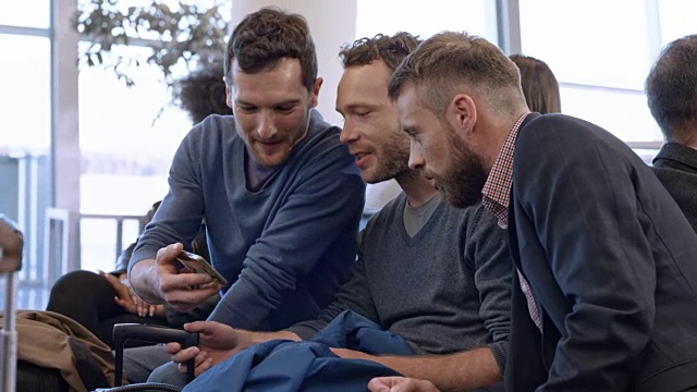 三个男人在机场等待登机时一边看手机一边笑视频素材
