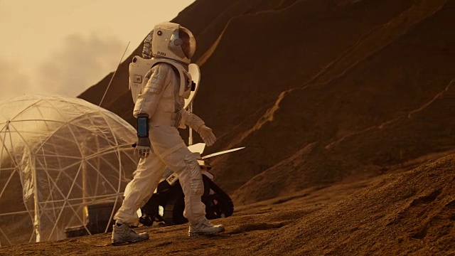 宇航员登上高山探索红色星球/火星。背景:他的基地和人工智能驱动的漫游车。未来殖民的概念。视频下载