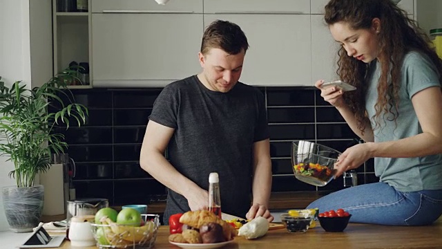 一对迷人的年轻夫妇在厨房里做饭，愉快地聊天。一名男子正在切蔬菜准备做沙拉，他的女朋友正在用智能手机拍照片发到社交媒体上视频素材