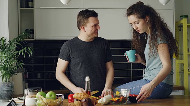 快乐的年轻夫妇做饭和聊天，而男人切蔬菜的早餐在厨房里在家。关系与家庭概念视频素材