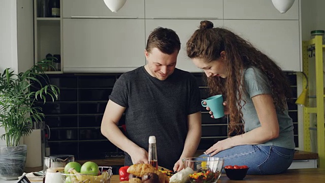 一对快乐的年轻夫妇在家里的厨房里做饭和聊天。帅哥一边喂女友一边切菜做早餐视频素材