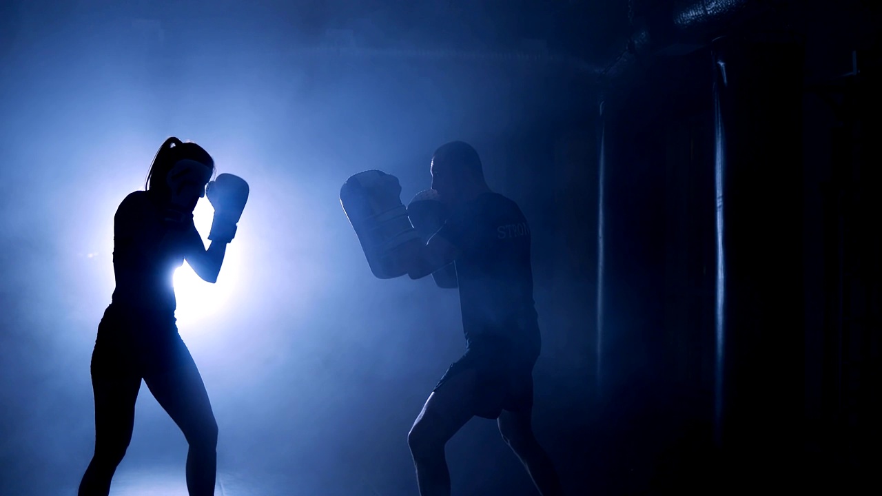 在烟雾缭绕的体育馆，一名女拳击手戴着拳击手套击打拳击袋的剪影视频下载