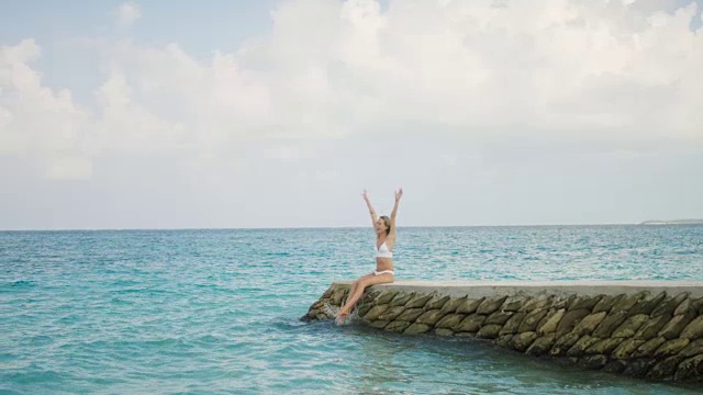 美丽的金发女郎穿着泳衣坐在码头上，举起手臂和微笑。快乐的人在度假，看海。视频素材