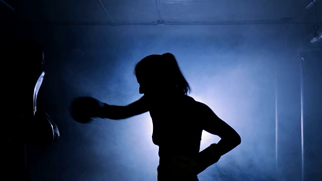 在烟雾缭绕的健身房里，一个年轻女子在拳击俱乐部教练的指导下训练视频素材