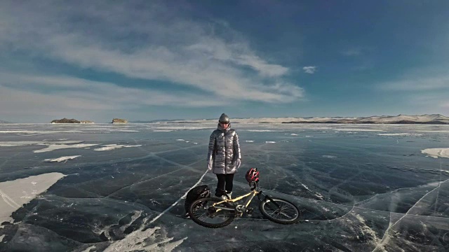 女人穿着运动器材。这个女孩穿着银色的羽绒服，背着自行车背包，戴着头盔。结冰的贝加尔湖的冰。自行车的轮胎上覆盖着特殊的尖钉。视频下载