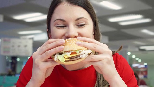 饥饿的女孩在美食广场吃汉堡。在快餐店里咬着芝士汉堡的女人视频素材