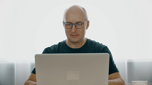一个戴眼镜的男人，在笔记本电脑后面工作，微笑着。男人露出了喜悦。一个穿着灰色t恤的快乐的人在笔记本电脑后面工作。成功和幸福的概念。视频下载