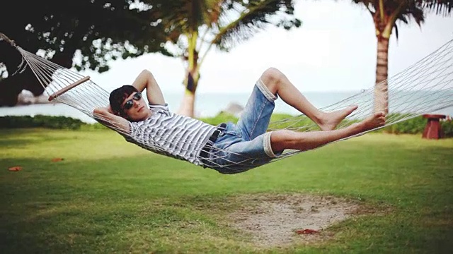 一个戴着墨镜的年轻人躺在美丽的热带海滩的吊床上休息。假期的概念。1920 x1080视频素材