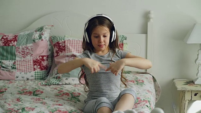 可爱的女孩戴着耳机坐在床上，在家里跳有趣的舞蹈视频素材