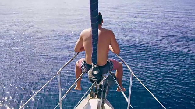 在阳光明媚的蓝色海洋上，一名男子正坐在帆船船头视频素材
