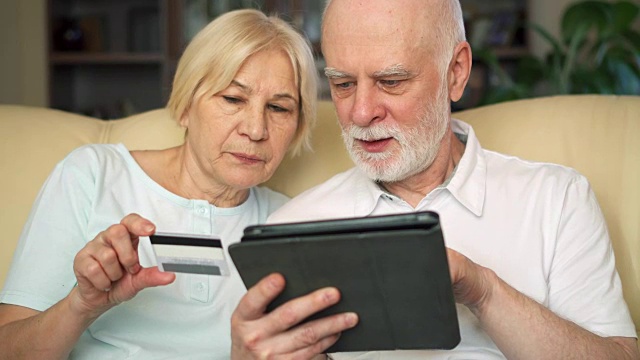 一对兴高采烈的好看的老夫妇坐在沙发上。在平板电脑上用信用卡在线购物视频素材
