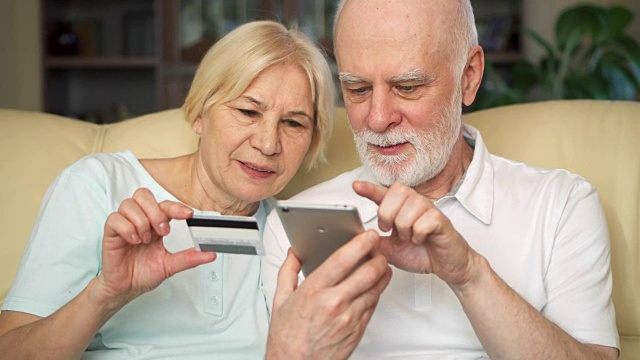 一对兴高采烈的好看的老夫妇坐在沙发上。在智能手机上用信用卡在线购物视频素材