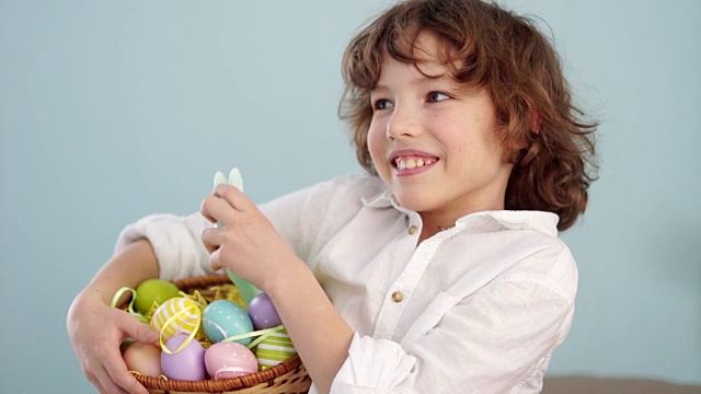 一个卷发的红头发的孩子，穿着一件白色衬衫，上面画着复活节兔子的图案，篮子里放着一套复活节彩蛋。一个男孩笑得很开心。缓慢的运动。视频下载
