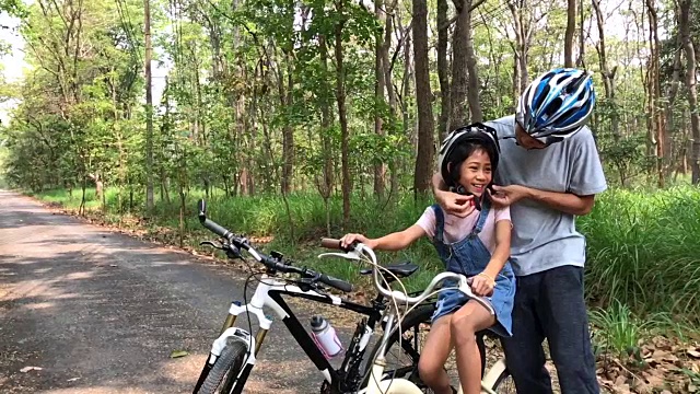 慢镜头快乐的爸爸和女儿骑着自行车在公园里给女儿戴上自行车头盔视频素材