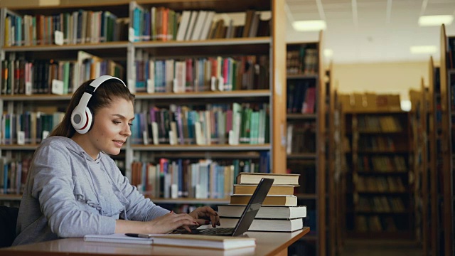 美丽积极的白人女学生与大耳机工作在宽敞的图书馆桌子前笔记本电脑。她微笑着发短信视频素材