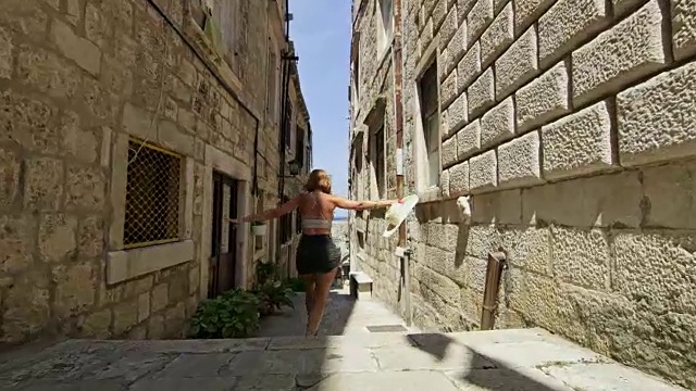 无忧无虑、顽皮的女游客在克罗地亚科尔库拉岛的阳光小巷里蹦蹦跳跳视频下载