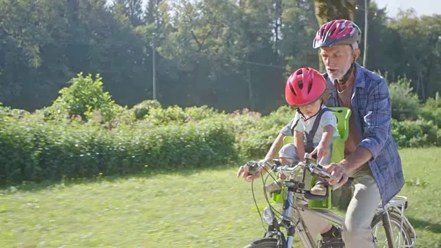 爷爷在阳光明媚的公园里骑着自行车，他蹒跚学步的孙子坐在前排按铃视频素材