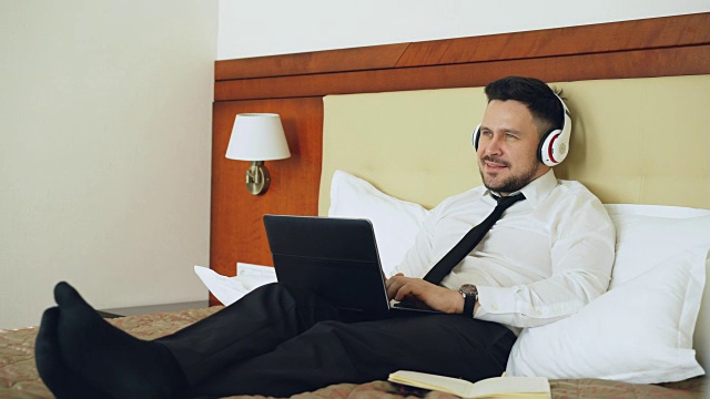 快乐的商人戴着耳机在笔记本电脑前工作，躺在酒店房间的床上微笑着听音乐。旅游、商务、人的理念视频素材