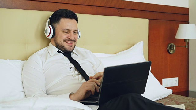 快乐的商人戴着耳机在笔记本电脑前工作，躺在酒店房间的床上微笑着听音乐。旅游、商务、人的理念视频素材