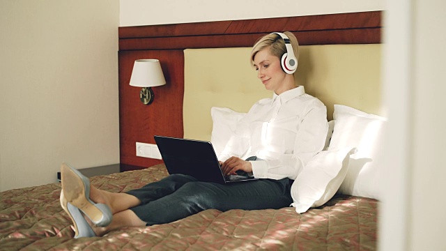 快乐的女商人戴着耳机在笔记本电脑前工作，躺在酒店房间的床上听着音乐微笑着跳舞。旅游、商务、人的理念视频素材