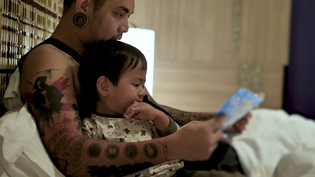 年轻的父亲和小儿子躺在家里的床上，在睡觉前阅读童话书视频下载