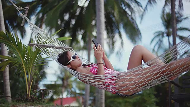 快乐年轻可爱的女孩躺在户外的秋千床上享受日光浴使用手机。1920 x1080视频素材