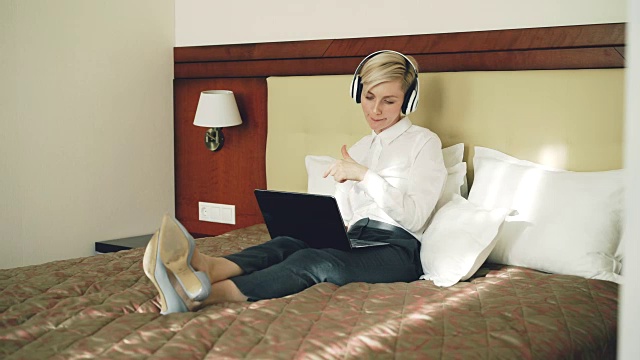 快乐的女商人戴着耳机听音乐，微笑着跳舞和唱歌，躺在酒店房间的床上用笔记本电脑。旅游、商务、人的理念视频素材