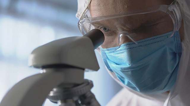 男实验室助理戴着防护口罩和眼镜，使用显微镜进行研究视频素材