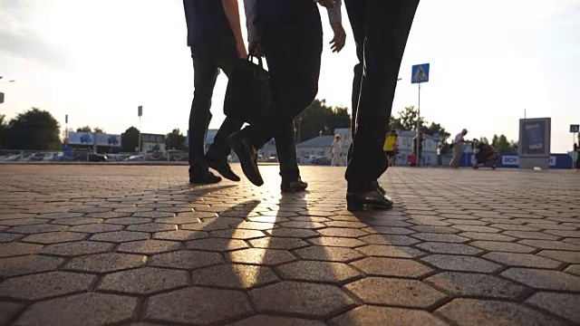 三个商人的脚走在机场附近，太阳耀斑的背景。一群面目全非的商人一起去码头。自信的人正在上班的路上。同事去户外。慢动作特写后视图视频素材