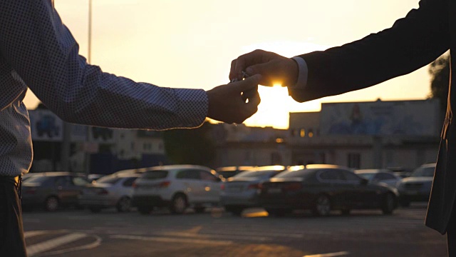 在太阳耀斑的背景下，男性将车钥匙交给商业伙伴。两个商人在户外握手。特写慢动作侧视图视频下载